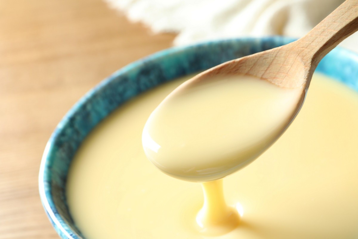 Spoon of pouring condensed milk over bowl on table, closeup with space for text. Dairy products