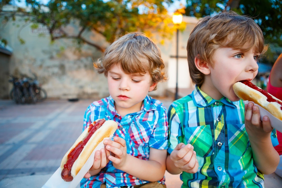 Two little kid boys eating hot dogs outdoors