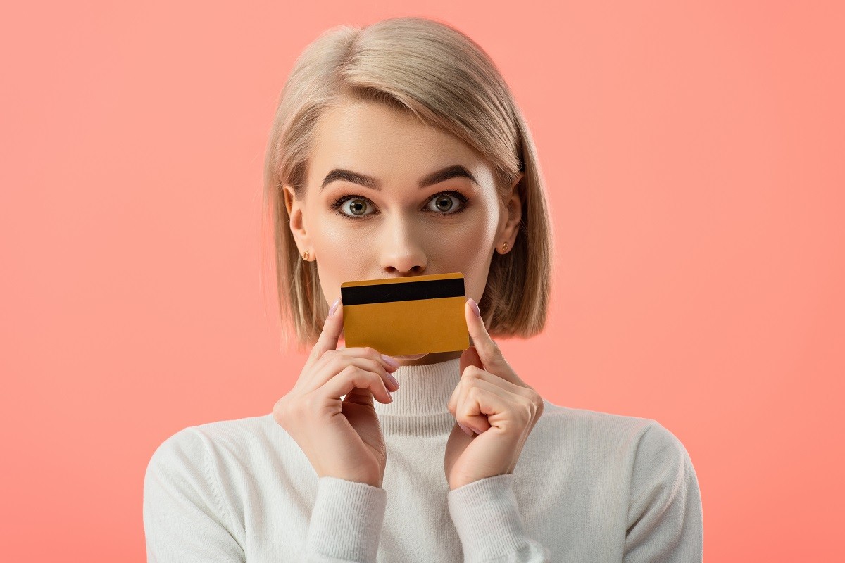 surprised blonde woman covering mouth with credit card isolated on pink