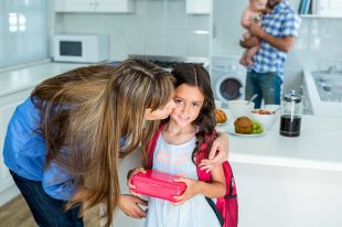 Mother kissing girl holding school lunch box with family in background at home