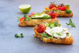 Variety of croissant sandwiches with grilled pepper, tomatoes, smoked salmon, turkey, avocado and arugula served with micro green on grey concrete background.