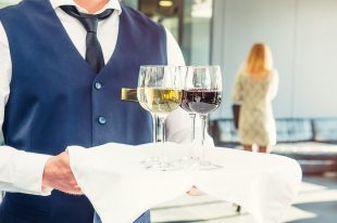 Professional waiter in uniform holding a tray with glasses of vine at business event. Catering or celebration concept. Service at corporate meeting, party, weddings. Selective focus, space for text.