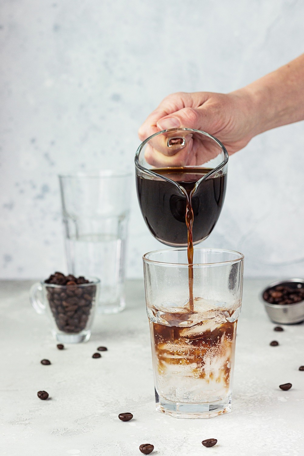 Pouring espresso into a tall glass with tonic and ice cubes, light grey stone background. Making tonic iced coffee. Refreshment summer drink.