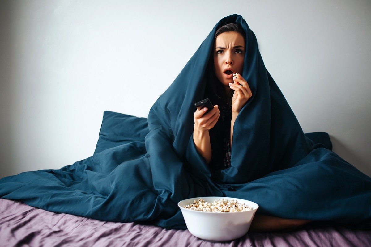 Young beautiful woman in morning bed at home. Watching scary movie alone in room. Covered with blanket. Eating popcorn and switching channels with remote control.