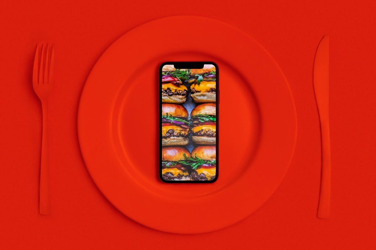Order food online, food delivery. Call and they’ll bring the food home. Smartphone with hamburgers on the screen, lying on a plate, a knife and fork. Red top View