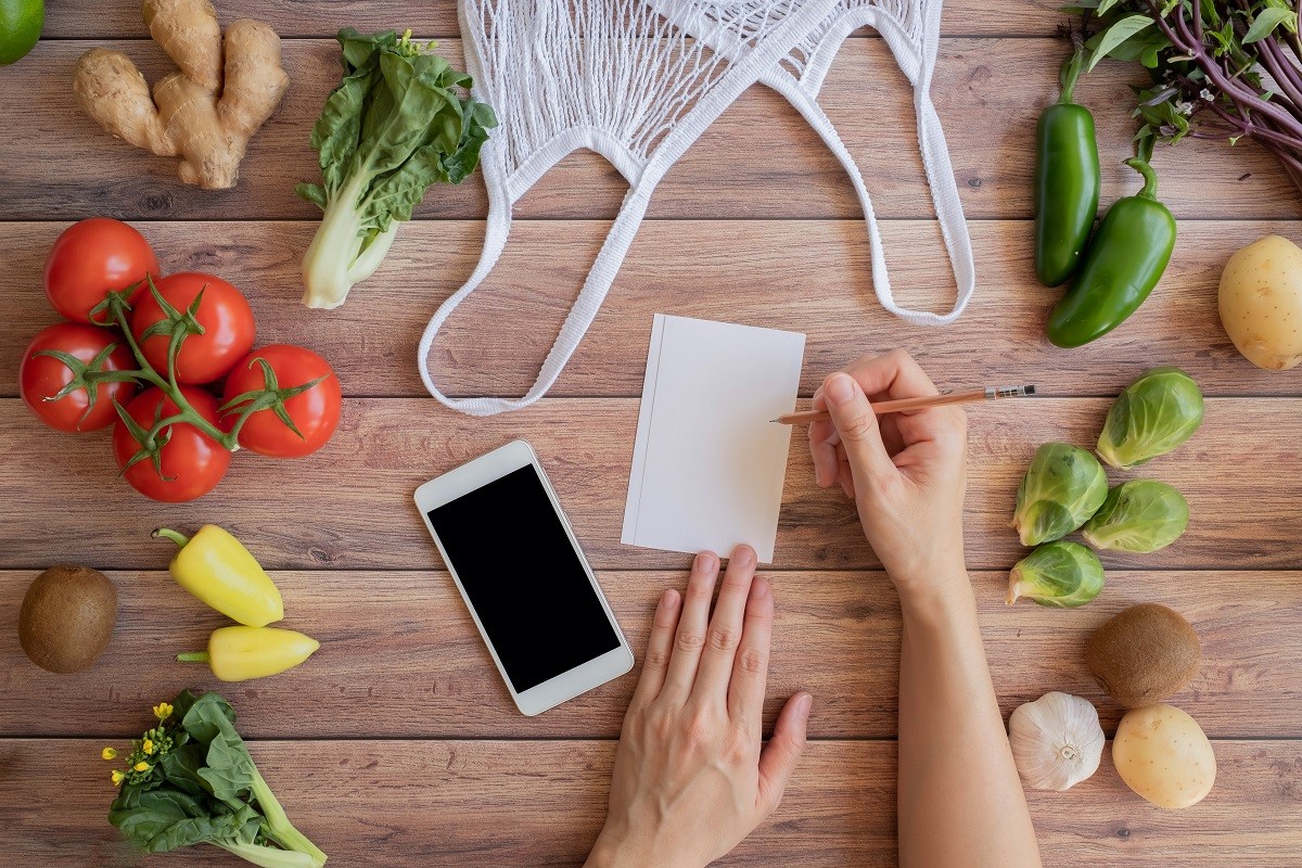 mobile phone and note list with the net eco bag and fresh vegetable on wood background. online grocery and organic farmer product shopping application. food and cooking recipe or nutrition counting.