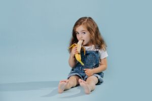 Little girl is eating banana, while sitting on the floor over blue background