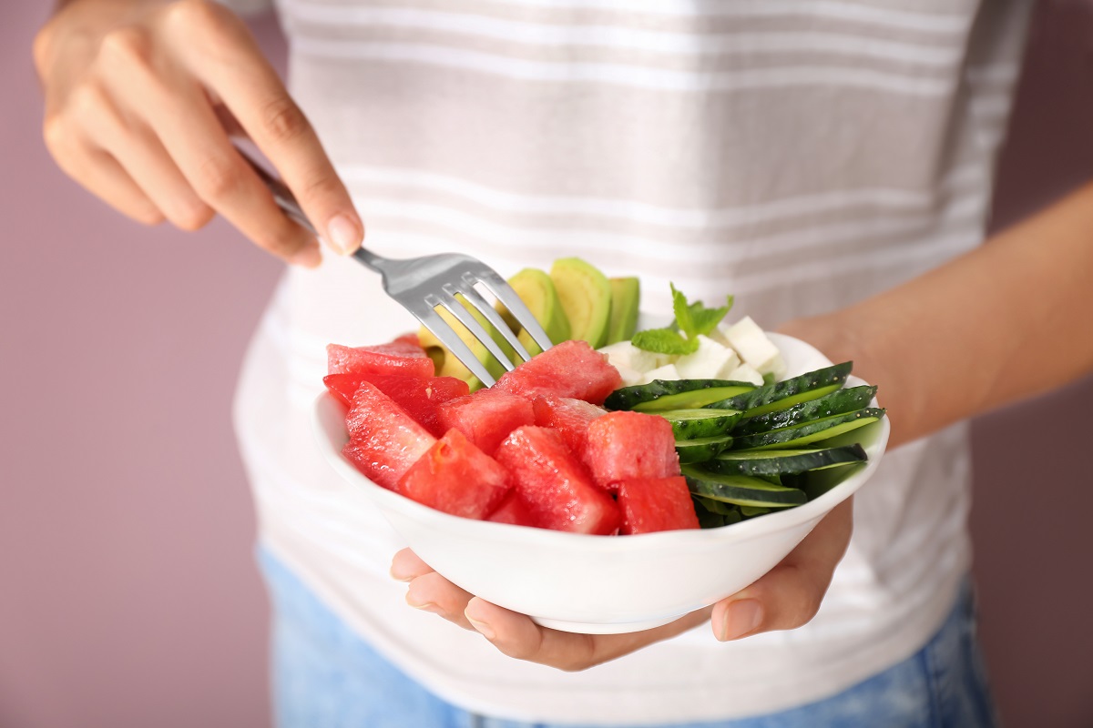 Woman eating tasty salad with watermelon, feta and vegetables on color background, closeup
