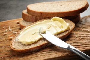 Spreading,Butter,Onto,Toast,With,Knife,On,Wooden,Board